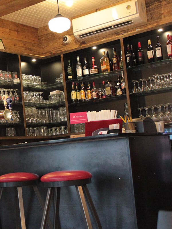 Deux tabourets devant un comptoir de bar avec à l'arrière des bouteilles d'alcools et verre de bières dans une étagère au mur.
