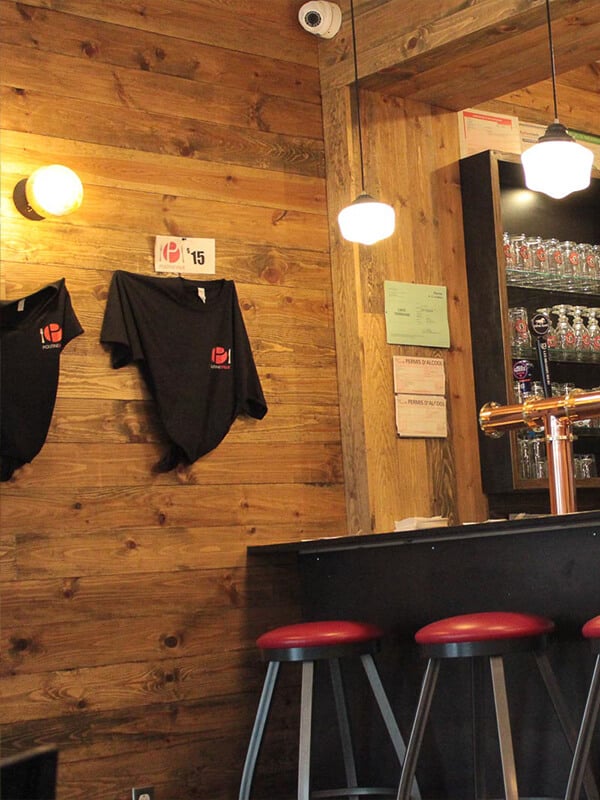 Deux tabourets devant un comptoir de bar avec une tour à bière en cuivre. Deux gilets noirs avec le logo de Poutineville à vendre sont affiché au mur.