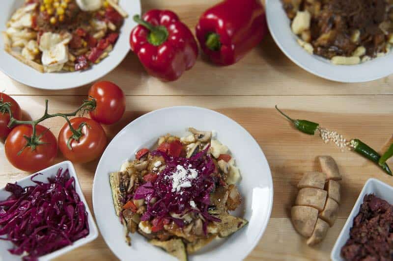 Poutine avec ingrédients frais servi dans une assiette entouré de légume frais (tomate en grappe, poivron rouge entier et patate) sur une grosse planche à découper en bois