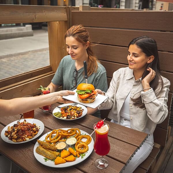 Trois amies assises à une terrasse du restaurant, mangeant une entrée, deux poutines et un burger accompagné de deux cocktails et une bière